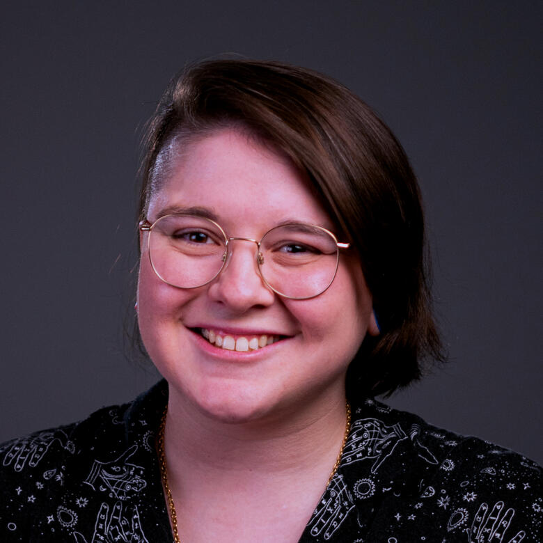 Photo by Tyrese Cortez A photograph of a person with a glasses. They are wearing a black button down shirt, with one side of their head shaved, and the other side with hair to their chin. They are smiling at the camera, facing head on.